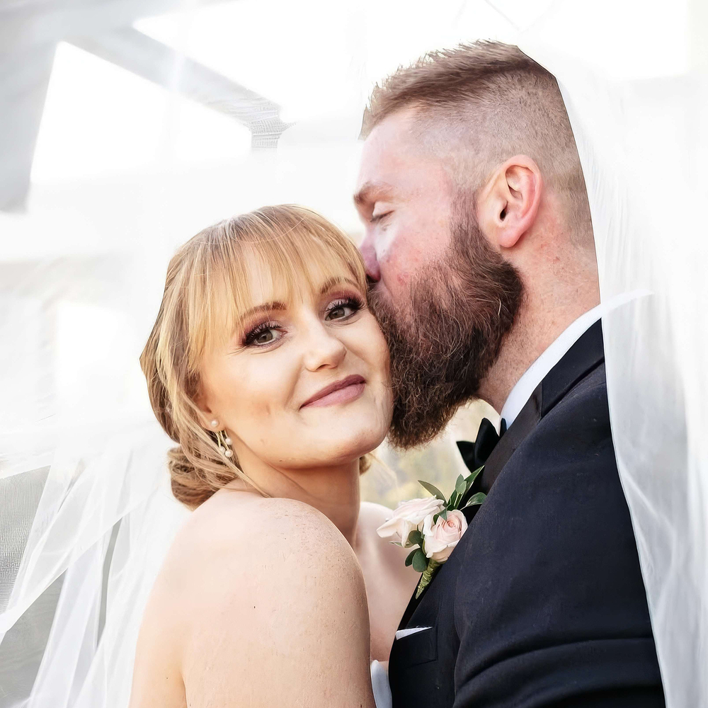 Bride smiling at camera while groom kisses cheek